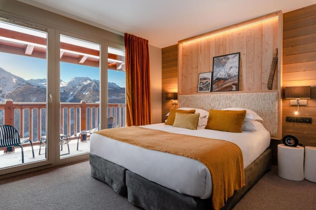 A bedroom with a large bed, a view of mountains, a wooden wall, and a balcony with chairs.