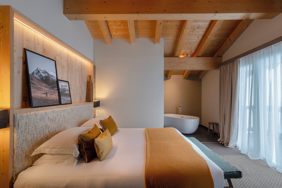 Ein modernes Hotelzimmer mit einem großen Bett, Holzboden, einem aufgehängten Bild, einer Badewanne und einem großen Fenster mit Vorhängen.