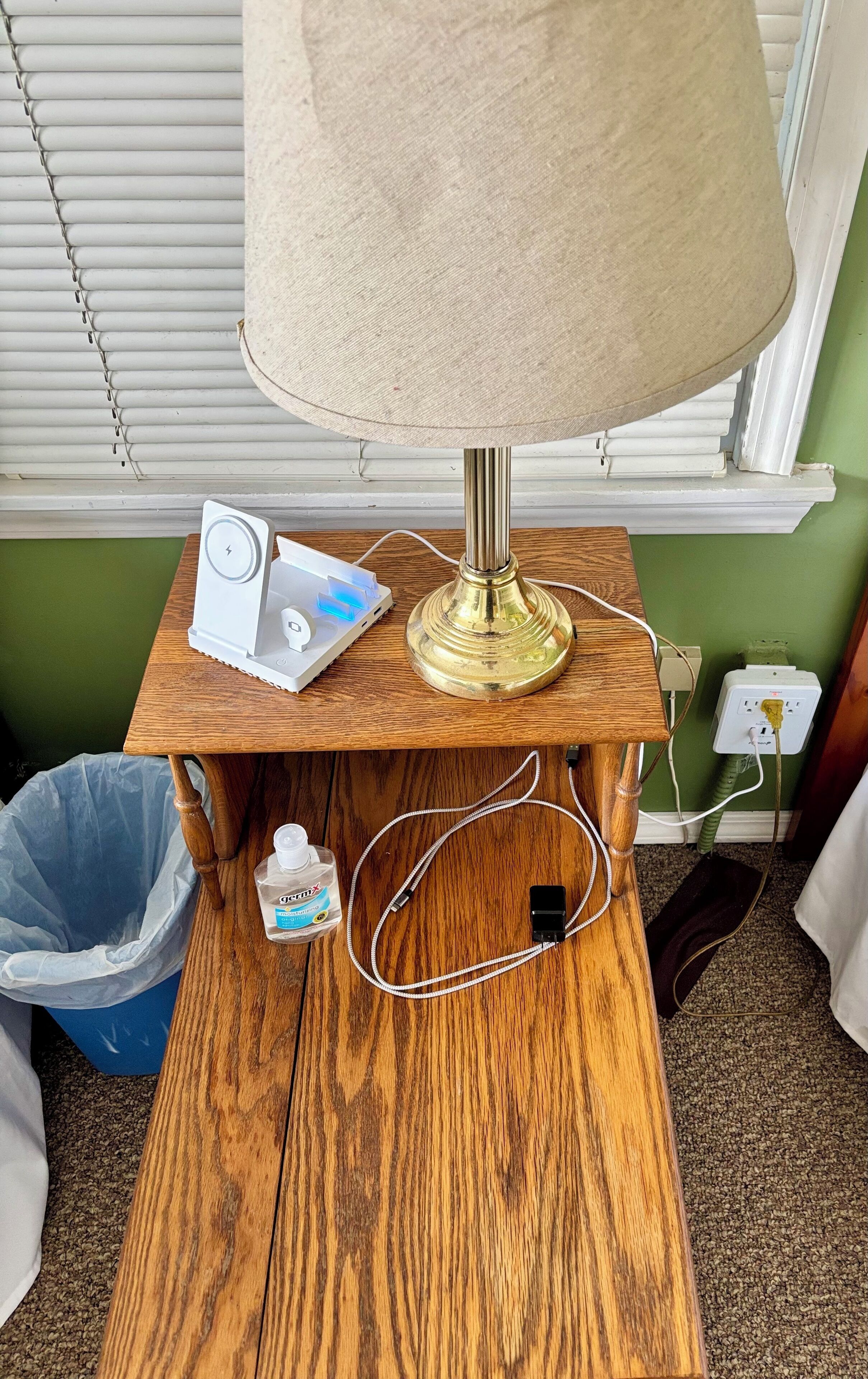 A wooden nightstand with a lamp, a white device, and a trash bin.