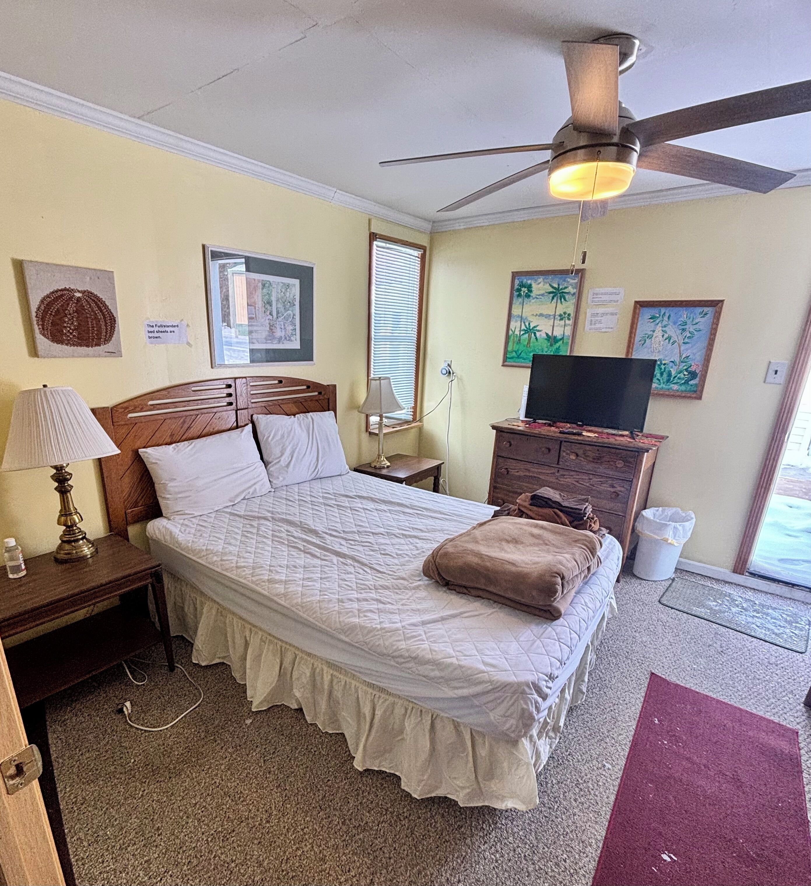Una habitación de hotel con cama, ventilador de techo, televisor y obras de arte en las paredes.