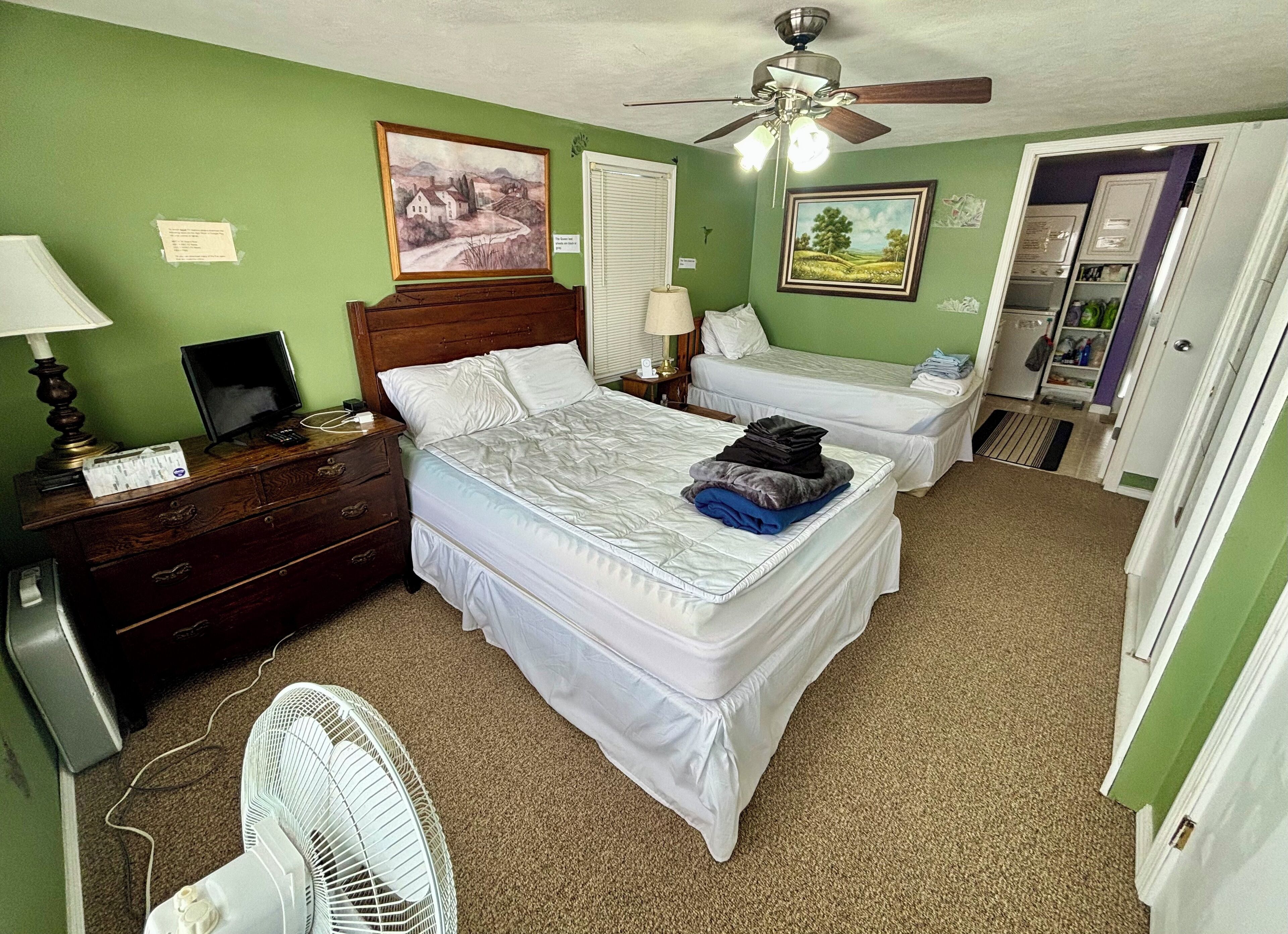 A bedroom with two beds, a ceiling fan, a desk with a lamp, and artwork on the walls.