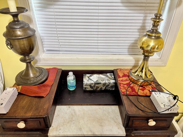 A room with a window with blinds, a table with a tissue box, a bottle of hand sanitizer, and a lamp on a stand. - The Wellington Cottage (Middlebury)
