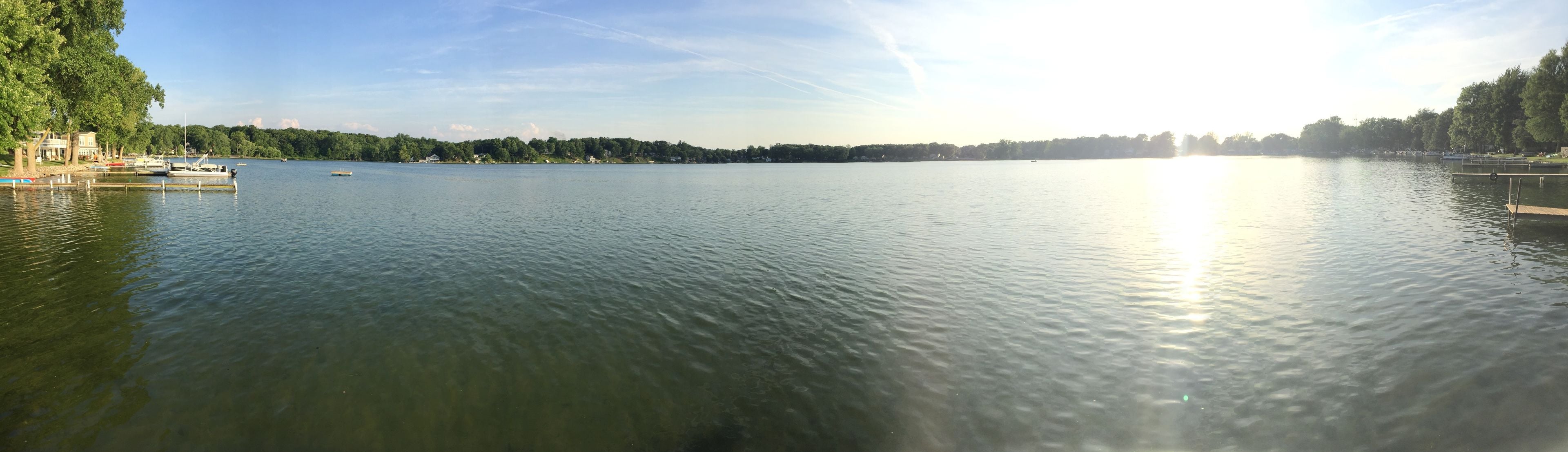 Un lago tranquilo con barcos amarrados a la izquierda, rodeado de árboles y un cielo despejado.