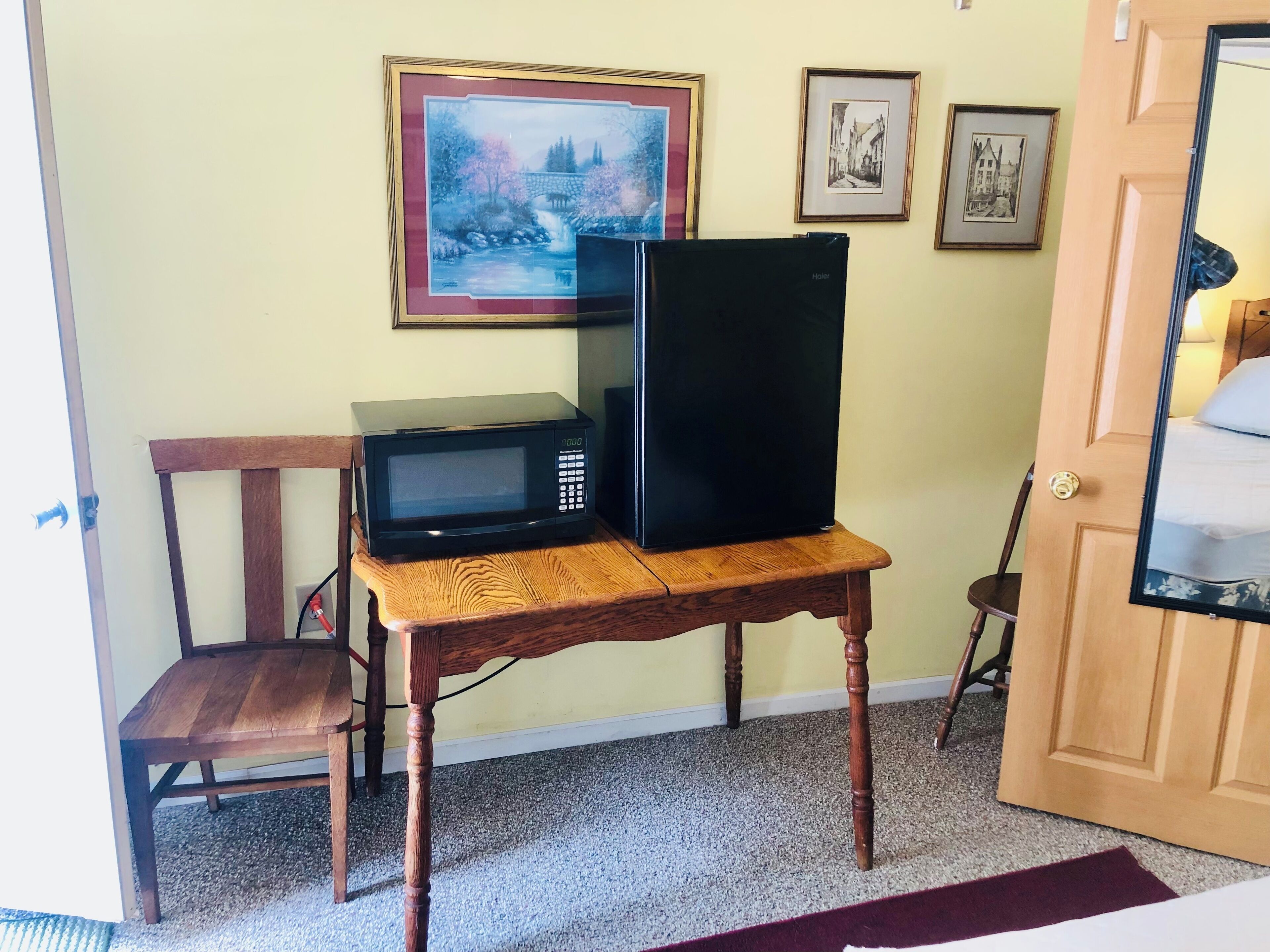 A room with a wooden table holding a microwave and a refrigerator, a chair, and a door with a mirror.