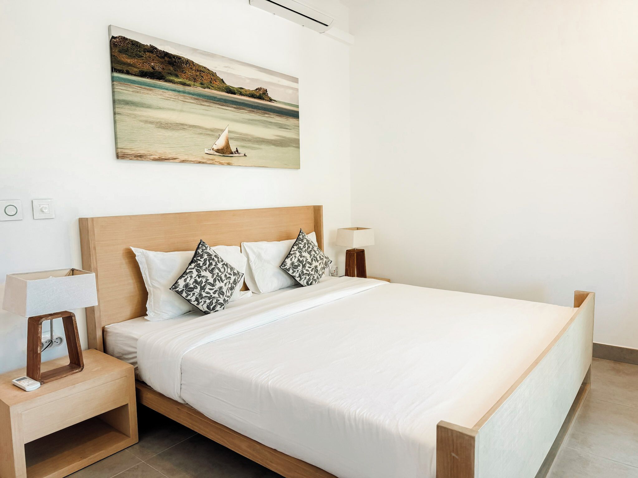 Une chambre à coucher avec un lit, deux oreillers, une table de chevet avec une lampe, un tableau accroché au mur et une petite table d’appoint.