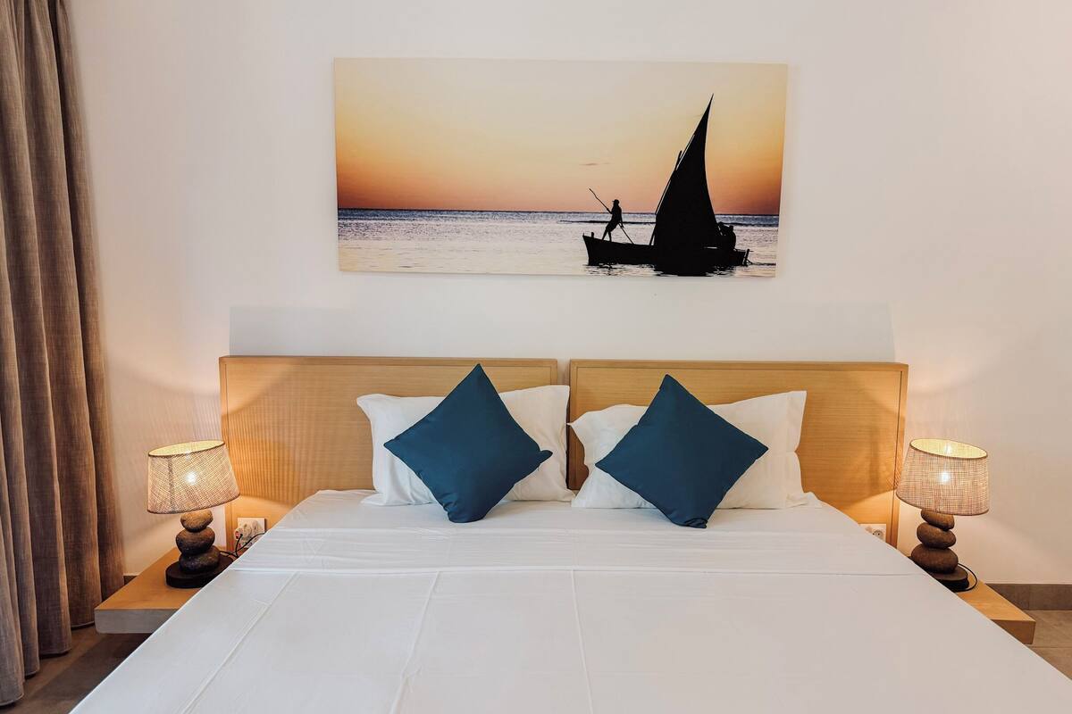 Une chambre d’hôtel avec un lit, deux oreillers, une table de chevet avec une lampe et un tableau représentant un voilier accroché au mur.