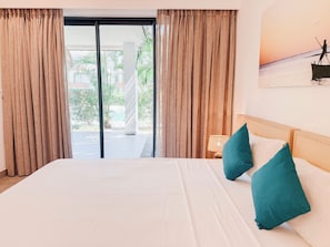 A hotel room with a bed, teal pillows, a nightstand with a lamp, and a view of a balcony and outdoor area.
