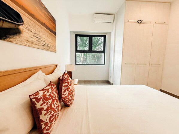 A bedroom with a bed, pillows, a nightstand, a window, an air conditioning unit, and a wardrobe.
