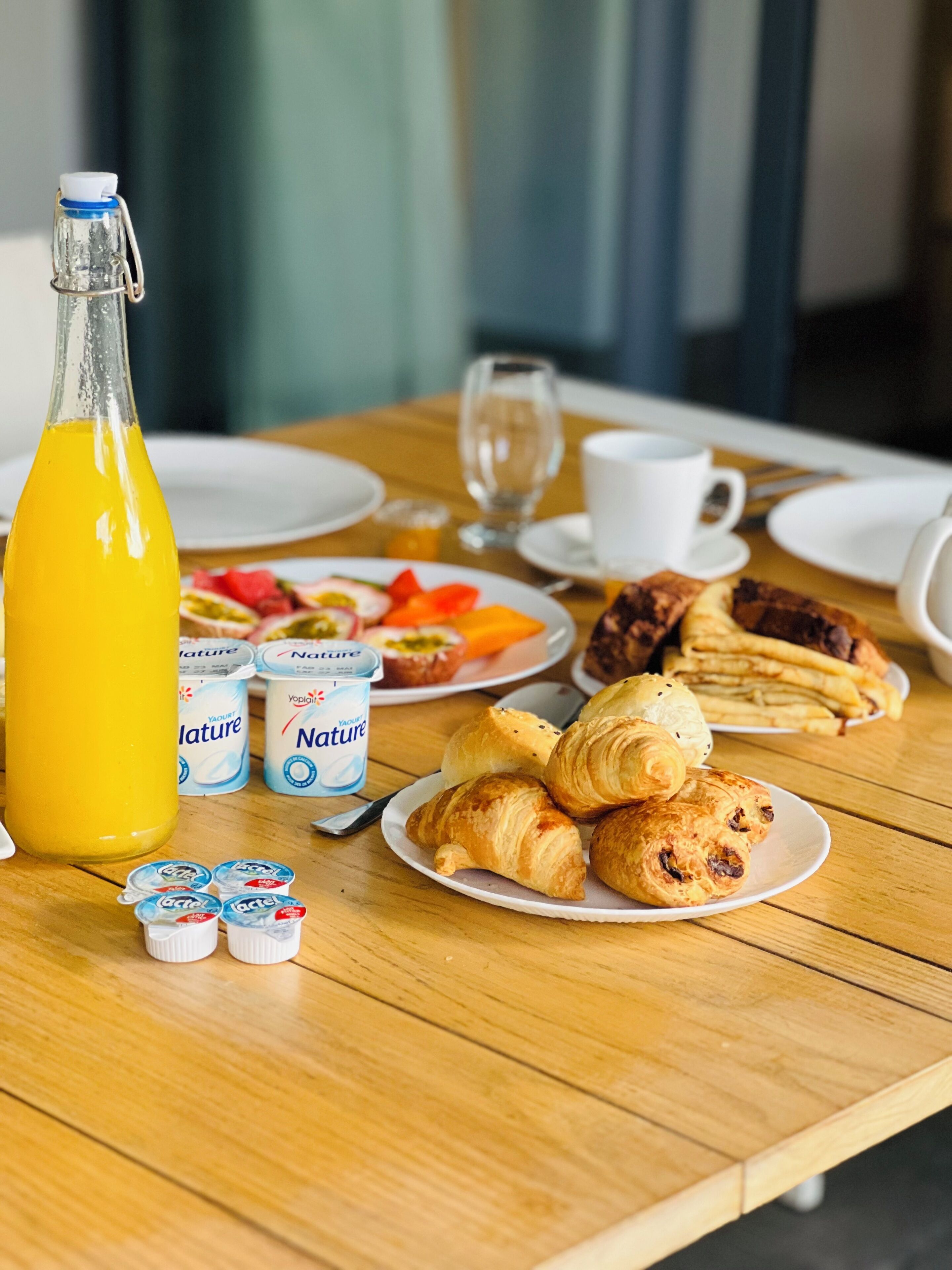 A breakfast table with a bottle of orange juice, yogurt cups, croissants, and a plate of fruit.