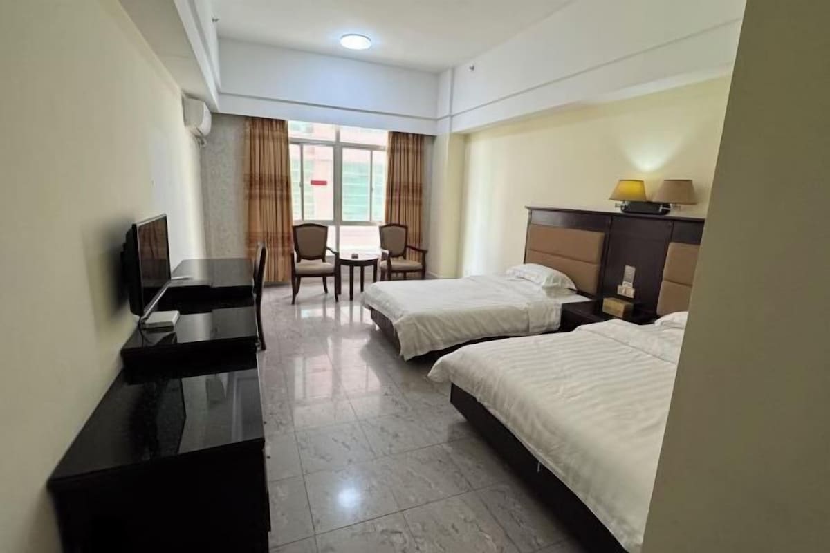 Habitación de hotel con dos camas, un escritorio con televisor de pantalla plana, una mesita con sillas y una ventana con cortinas.