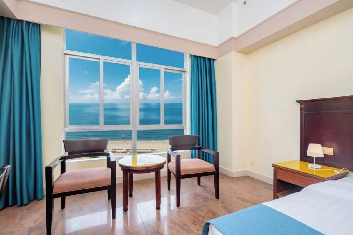 Habitación de hotel con un ventanal grande, una cama, dos sillas, una mesita y una mesita de noche con lámpara.