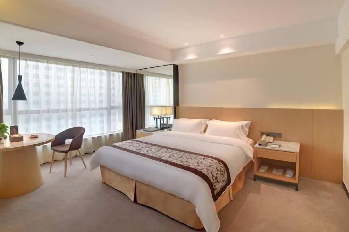 A hotel room with a large bed, bedside tables, a desk with a chair, and a large window with curtains.
