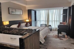 Habitación de hotel con cama, escritorio, silla, ventana grande con cortinas, baño con bañera y un cuadro en la pared.