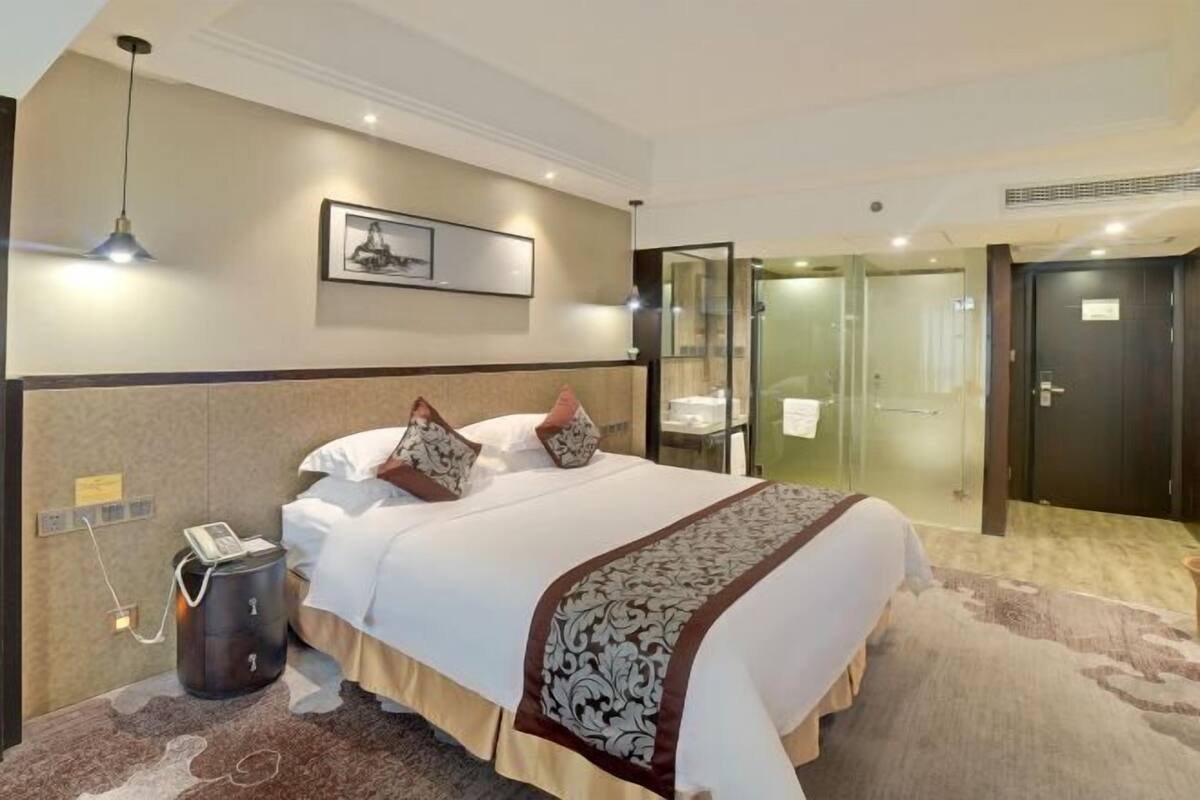 A hotel room with a large bed, a bedside telephone, a framed picture on the wall, and a glass-enclosed shower area.