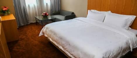 A hotel room with a bed, a small table with flowers, a chair, and a wooden desk.