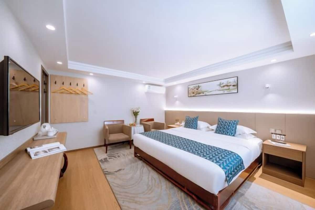 A hotel room with a large bed, a desk with a chair, a small table with a vase, and a painting on the wall.