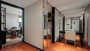 Habitació d'hotel moderna amb un separador de vidre, zona de menjador amb taula i cadires, i bany visible amb mirall i suport per tovalloles.