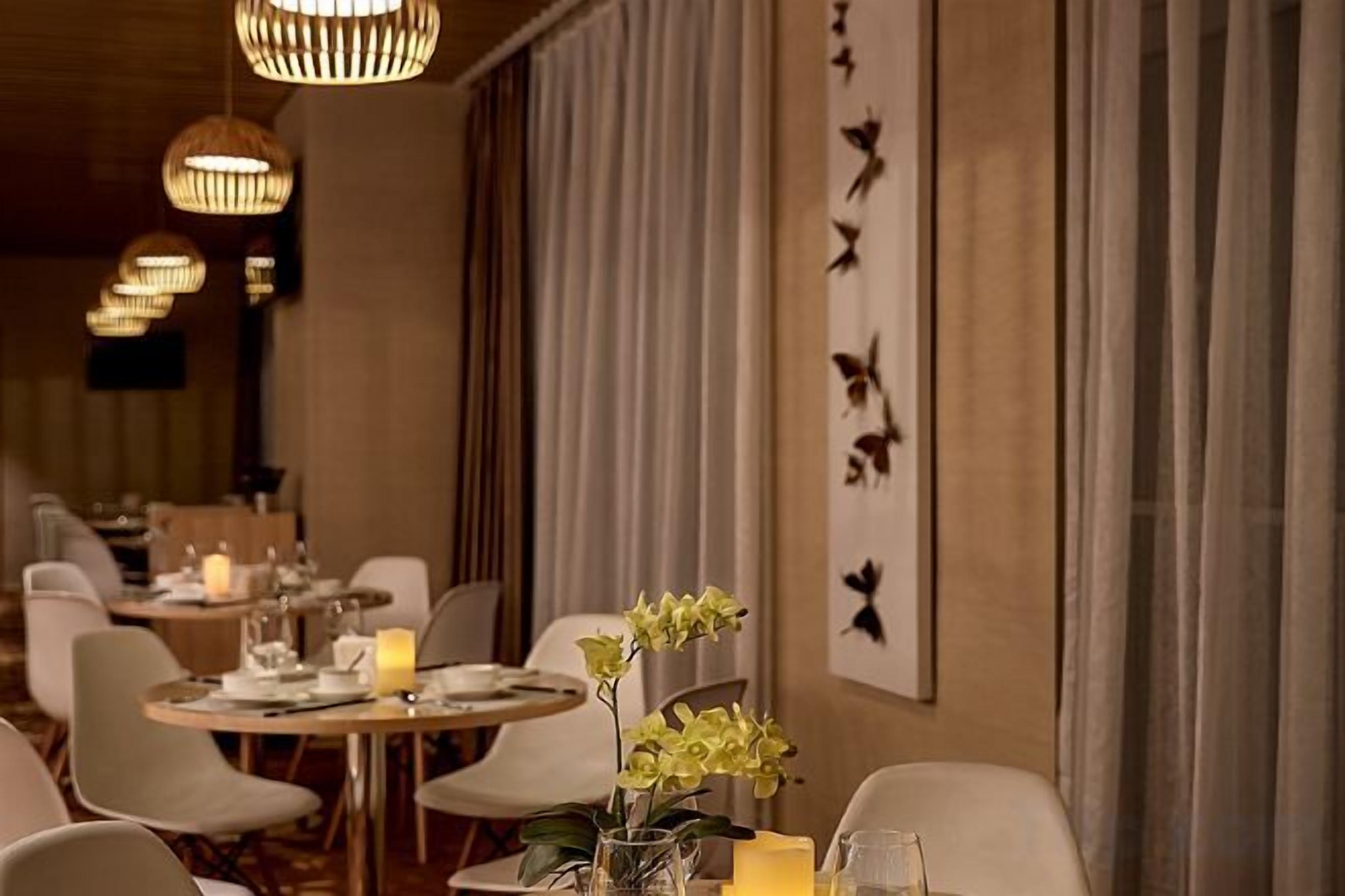 Restaurante com mesas redondas, cadeiras, velas e uma parede com silhuetas de pássaros.