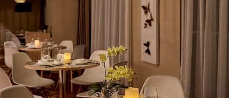 Restaurante com mesas redondas, cadeiras, velas e uma parede com silhuetas de pássaros.