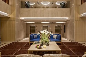 Un vestíbulo con techo alto, un área de estar central con sofás azules, una mesa con un jarrón de flores y una araña.