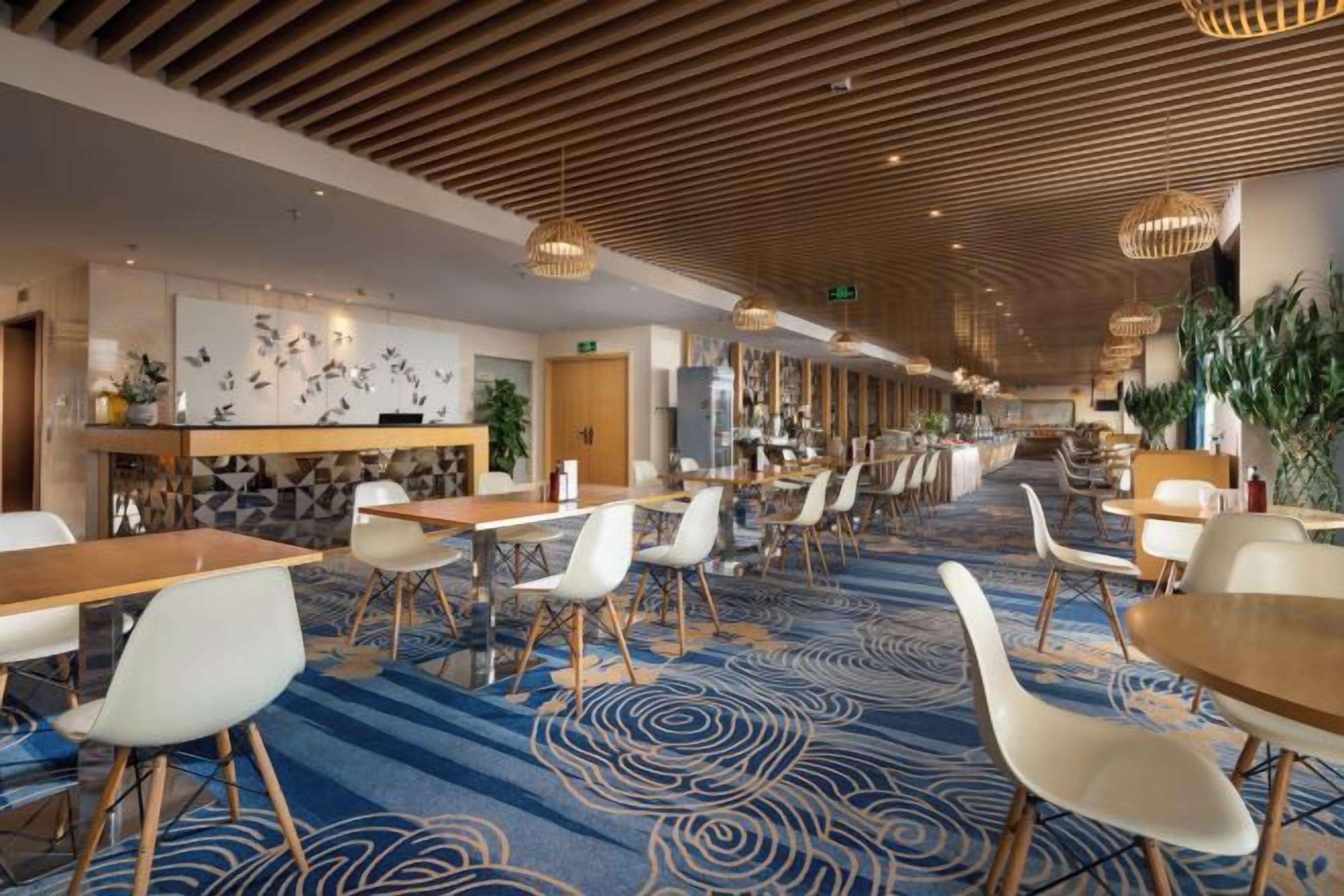A hotel lobby with a long corridor lined with tables and chairs, a patterned carpet, and a wooden ceiling.