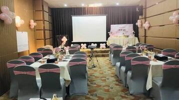 Un salón de conferencias con filas de mesas, sillas y un escenario con una pantalla de presentación.