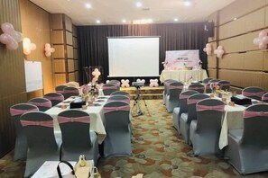 Un salón de conferencias con filas de mesas, sillas y un escenario con una pantalla de presentación.