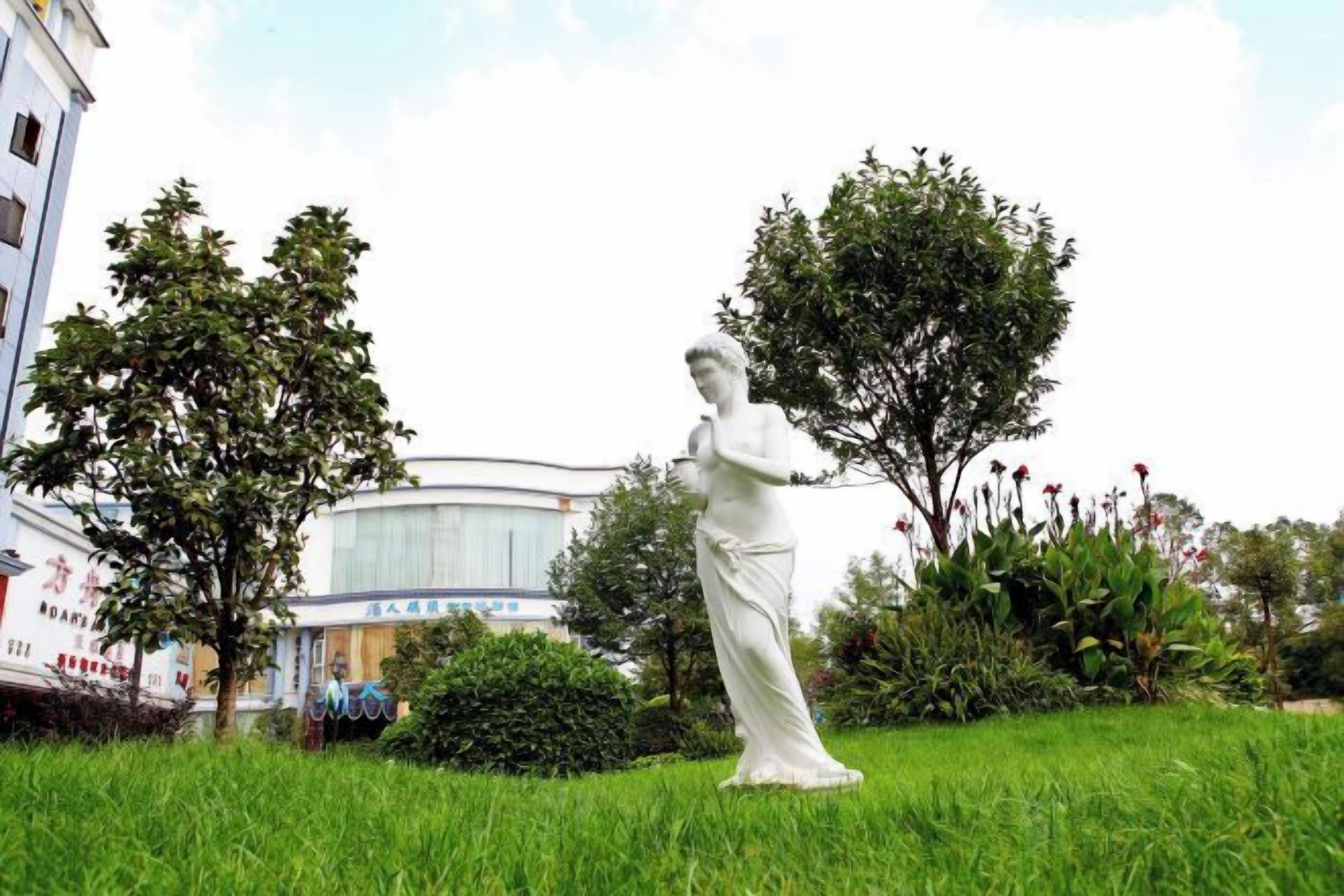 Eine Statue in einer grünen Parklandschaft, im Hintergrund sind Gebäude zu sehen.