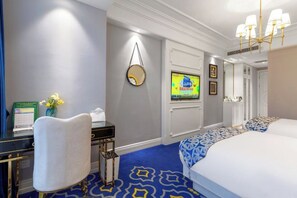Habitación de hotel con cama, escritorio, silla, televisor, espejo y alfombra estampada.