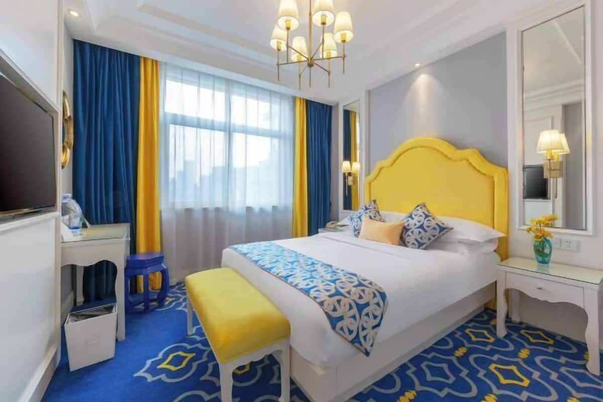 A hotel room with a large bed, a desk, a chair, a bench, a TV, and a window with curtains.