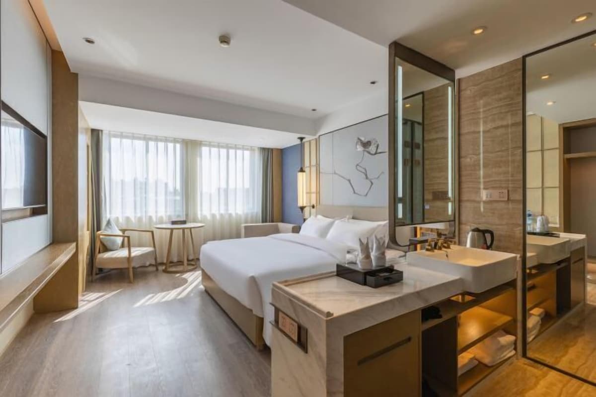 A modern hotel room with a large bed, bedside tables, a desk, and a bathroom with a mirror and sink.