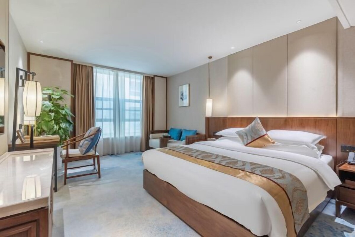 Hotellihuone, jossa on suuri sänky, työpöytä, tuoli, yöpöytä, lamppu ja suuri ikkuna, jossa on verhot.