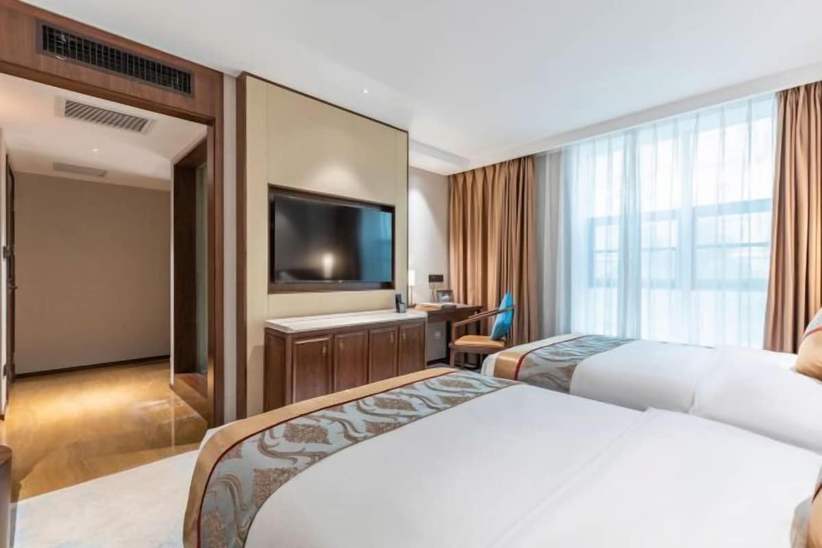 A hotel room with two beds, a TV, a desk, and a large window with curtains.