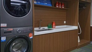 A compact laundry room with a washing machine, a sink, and a shelf with bottles. - Piaget Hotel (Anqing)