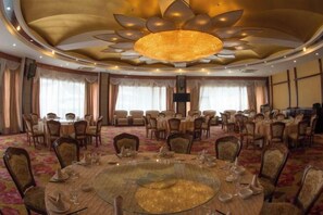 Un salón de banquetes con mesas redondas, sillas, una gran lámpara de araña y suelo alfombrado.