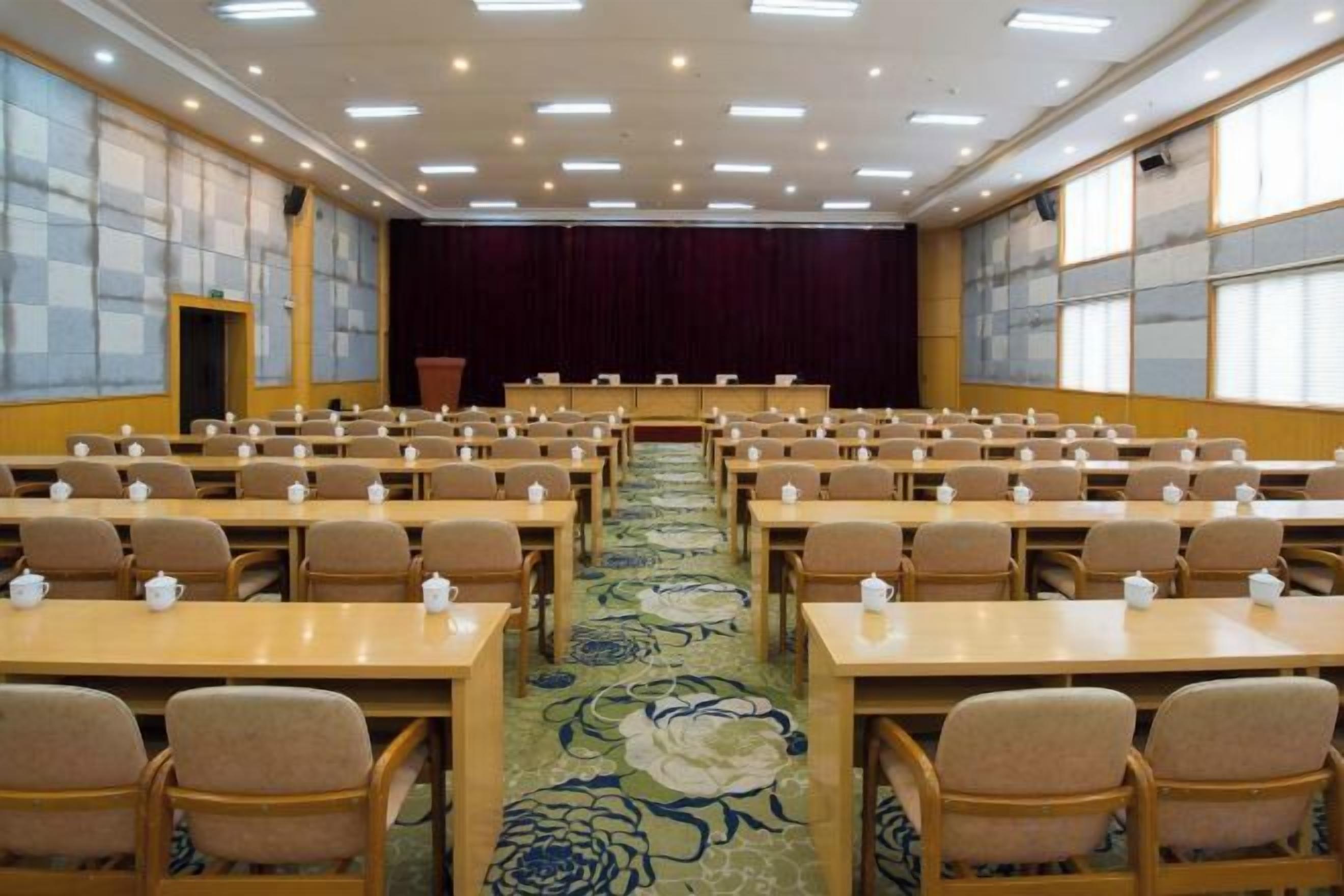 Un salón de conferencias con filas de mesas y sillas, una alfombra con estampado floral y un escenario con cortina.