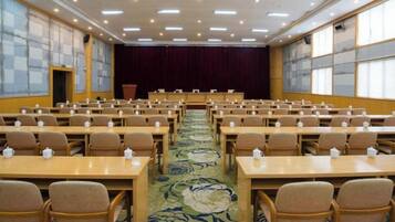 Un salón de conferencias con filas de mesas y sillas, una alfombra con estampado floral y un escenario con cortina.