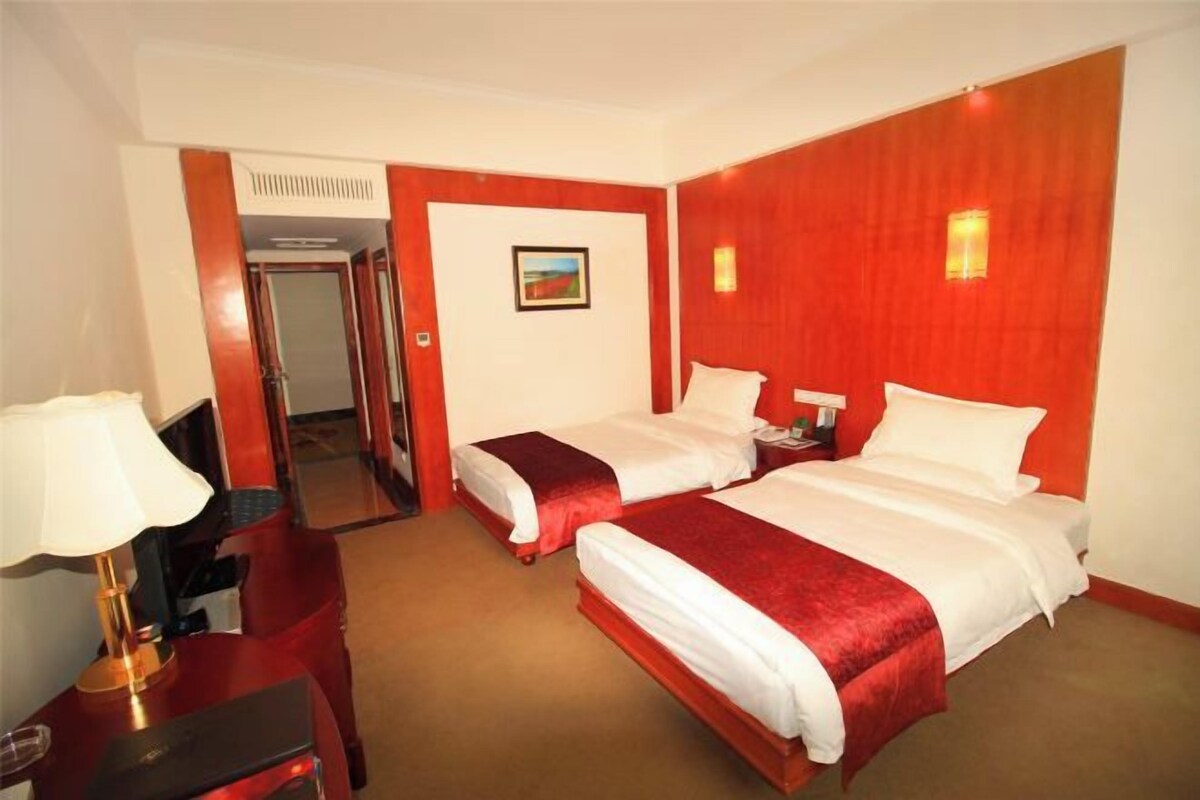 A hotel room with two beds, a desk with a lamp, a chair, and a door leading to another room.