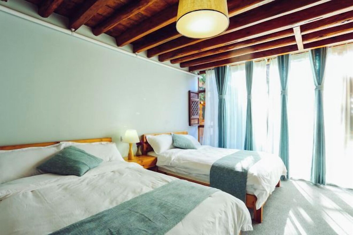 A room with two beds, wooden beams on the ceiling, a hanging light fixture, and a window with curtains.