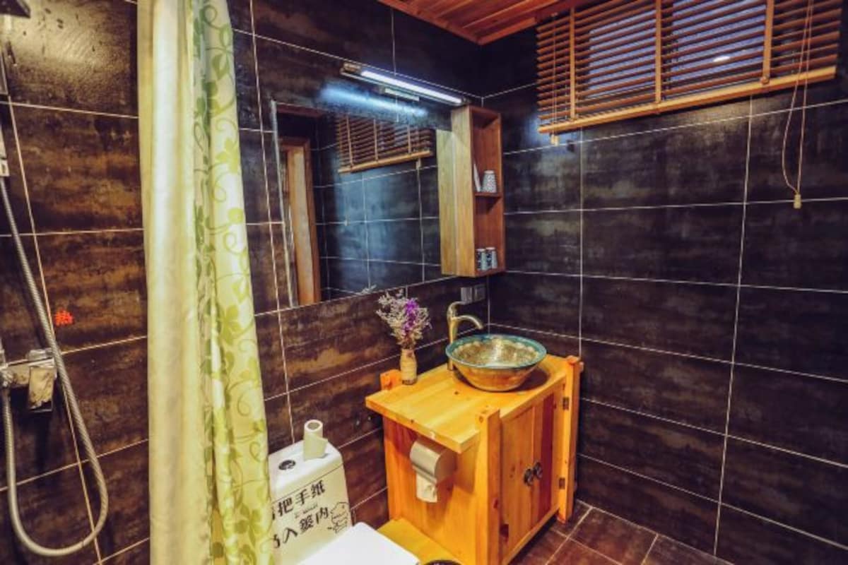 A bathroom with dark tiled walls, a wooden cabinet with a glass bowl sink, a shower with a curtain, and a mirror above the sink.