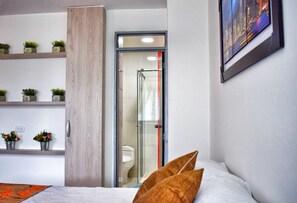 A modern bathroom with a glass shower enclosure, a toilet, and a sink. There are shelves with potted plants and a bed with a pillow. - Apartahotel Casa del Lido (Cali)