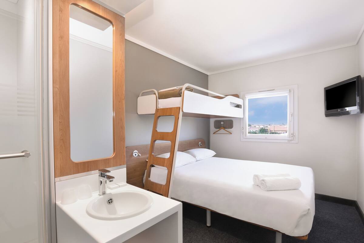 Ein Hotelzimmer mit Etagenbett, Badezimmer mit Waschbecken und Spiegel sowie einem an der Wand befestigten Fernseher.