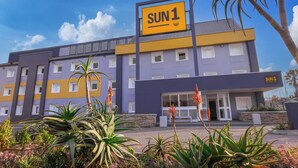 A hotel with a 'SUN1' sign, featuring a modern design with purple and yellow colors, multiple windows, and a well-maintained garden with various plants. - SUN1 Milnerton (Cape Town)