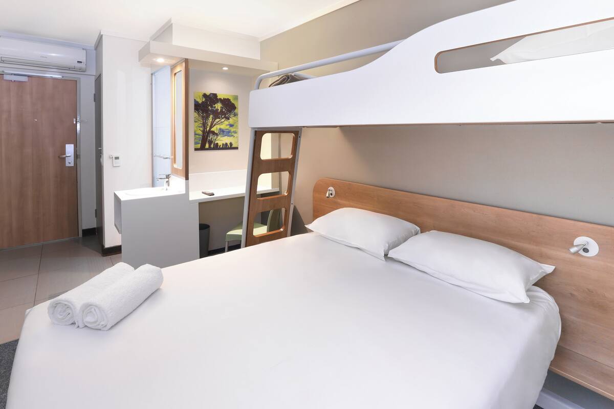 Ein Etagenbett mit Holz-Kopfteil, ein weißes Badezimmer mit Waschbecken und Spiegel sowie ein Bild an der Wand.