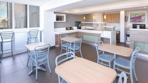A modern hotel dining area with multiple tables and chairs, a kitchen area with a refrigerator and coffee machine, and a large window providing natural light. - SUN1 Milnerton (Cape Town)