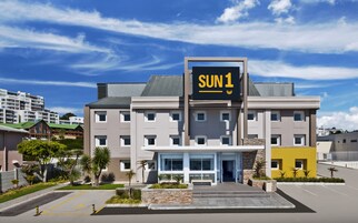 Un hotel moderno con un llamativo letrero 'SUN 1', que presenta una entrada central con fachada de vidrio, un estacionamiento y zonas verdes circundantes.