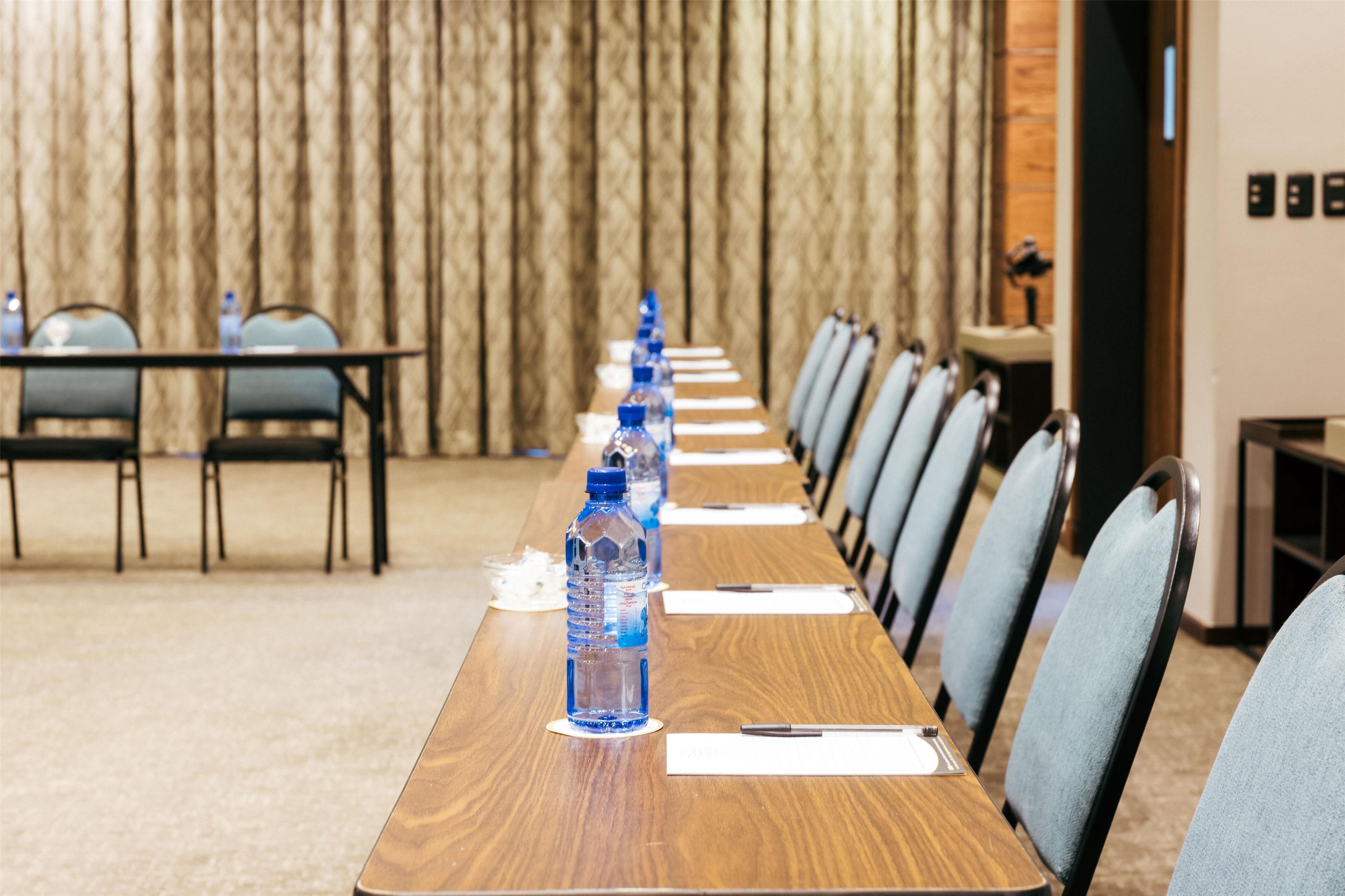 A conference room with a long table, chairs, water bottles, and a wooden backdrop.