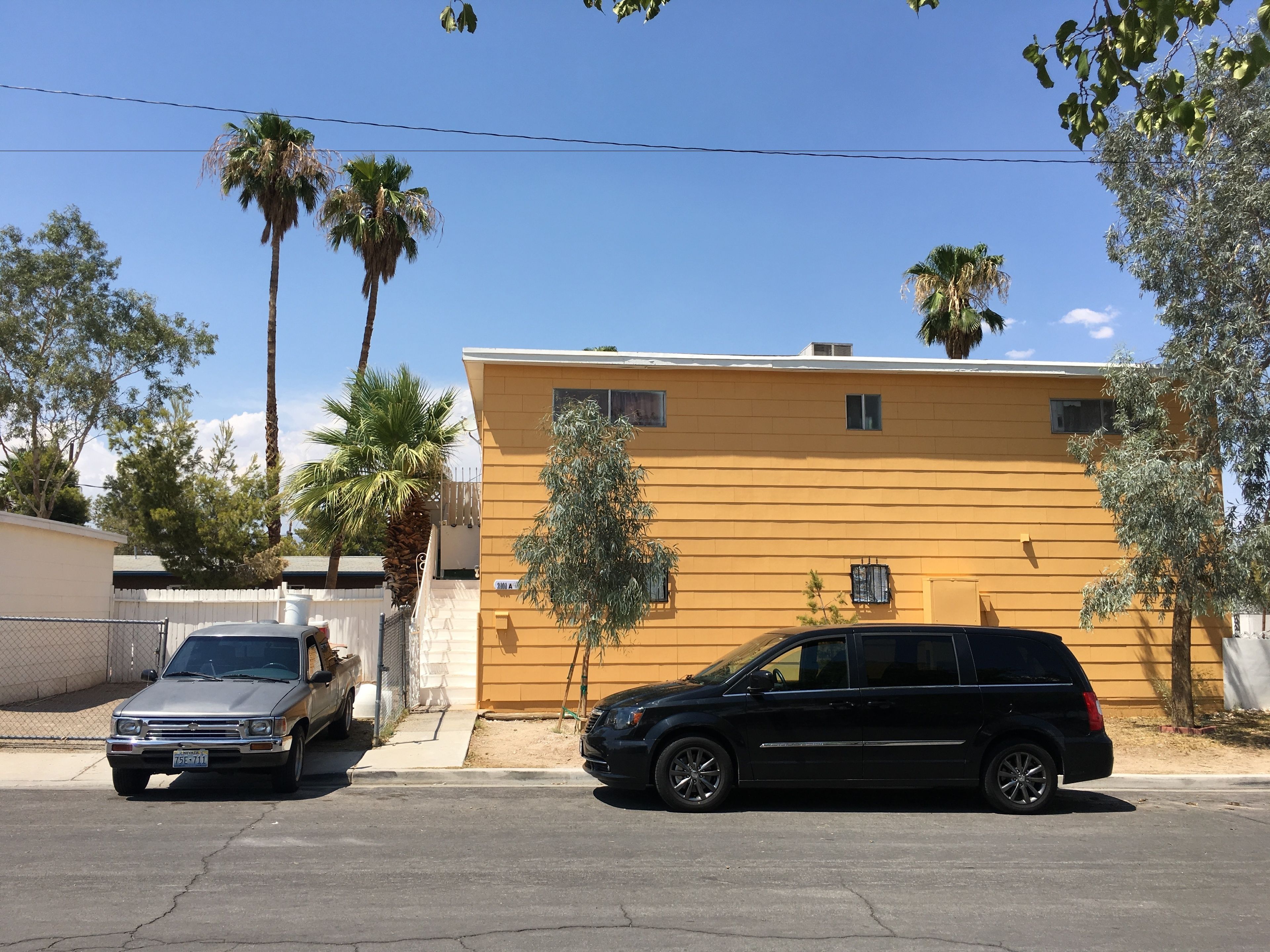 Un edificio amarillo con una entrada protegida por una reja, dos coches aparcados y palmeras.
