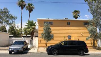 Un edificio amarillo con una entrada protegida por una reja, dos coches aparcados y palmeras.