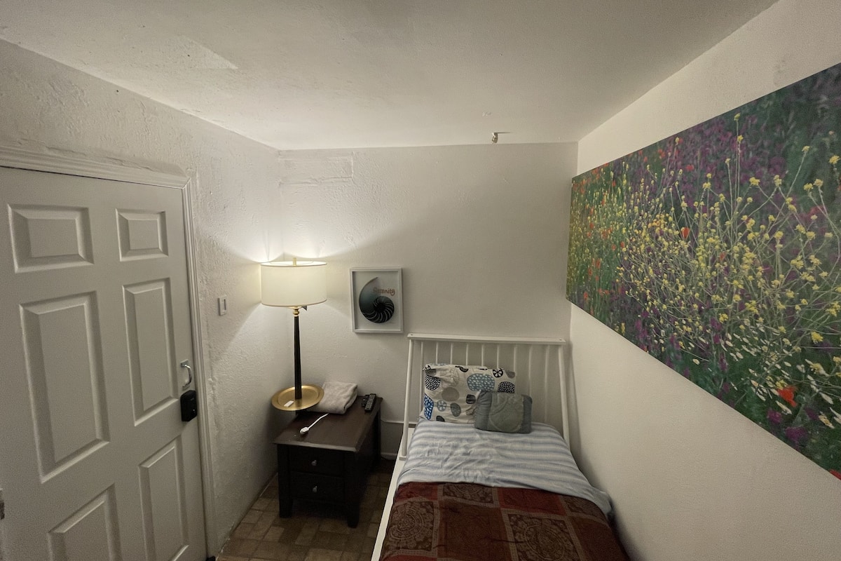 Un dormitorio con una cama, una mesita de noche, una lámpara, un radiador y un cuadro con motivos florales en la pared.